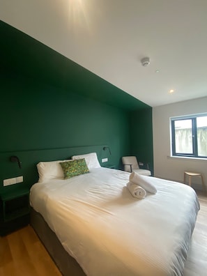 Habitació d'hotel amb un llit gran, una paret verda decorativa, una finestra amb cortines i una tauleta de nit amb una làmpada.