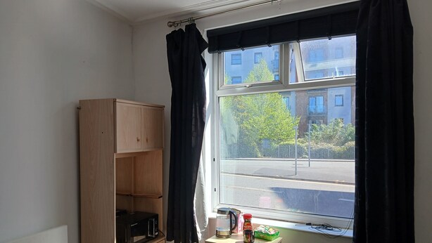 A room with a window, a wooden cabinet, a stool, a bed, and a small table with a kettle and a mug.