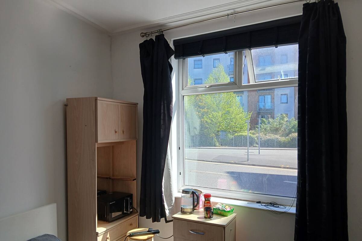A room with a window, a wooden cabinet, a stool, a bed, and a small table with a kettle and a mug.