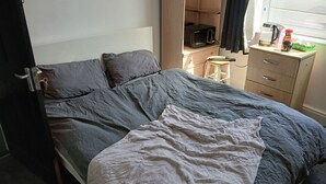 A bedroom with a bed, a nightstand, a microwave, a kettle, and a window with curtains.