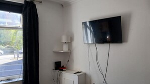A room with a TV mounted on the wall, a window with a curtain, a small white cabinet, and a brown sofa.