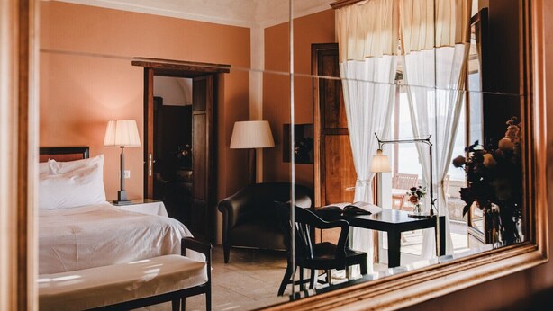 Een hotelkamer met een bed, een bureau met stoel, een spiegel en een raam met gordijnen.