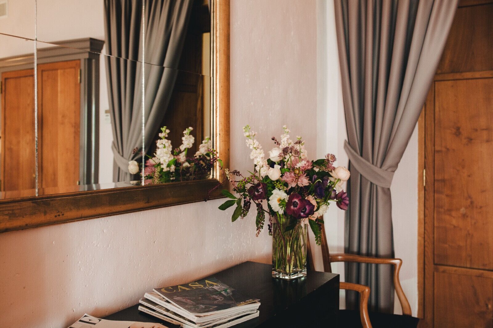 Ein Zimmer mit einem Holzkabinett, einem Spiegel der ein Fenster mit Vorhängen reflektiert, einer Vase mit Blumen und einem Stapel Zeitschriften auf einem Tisch.