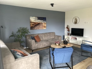 A living room with a grey sofa, a blue armchair, a coffee table, a TV on a white stand, and a large painting on the wall.