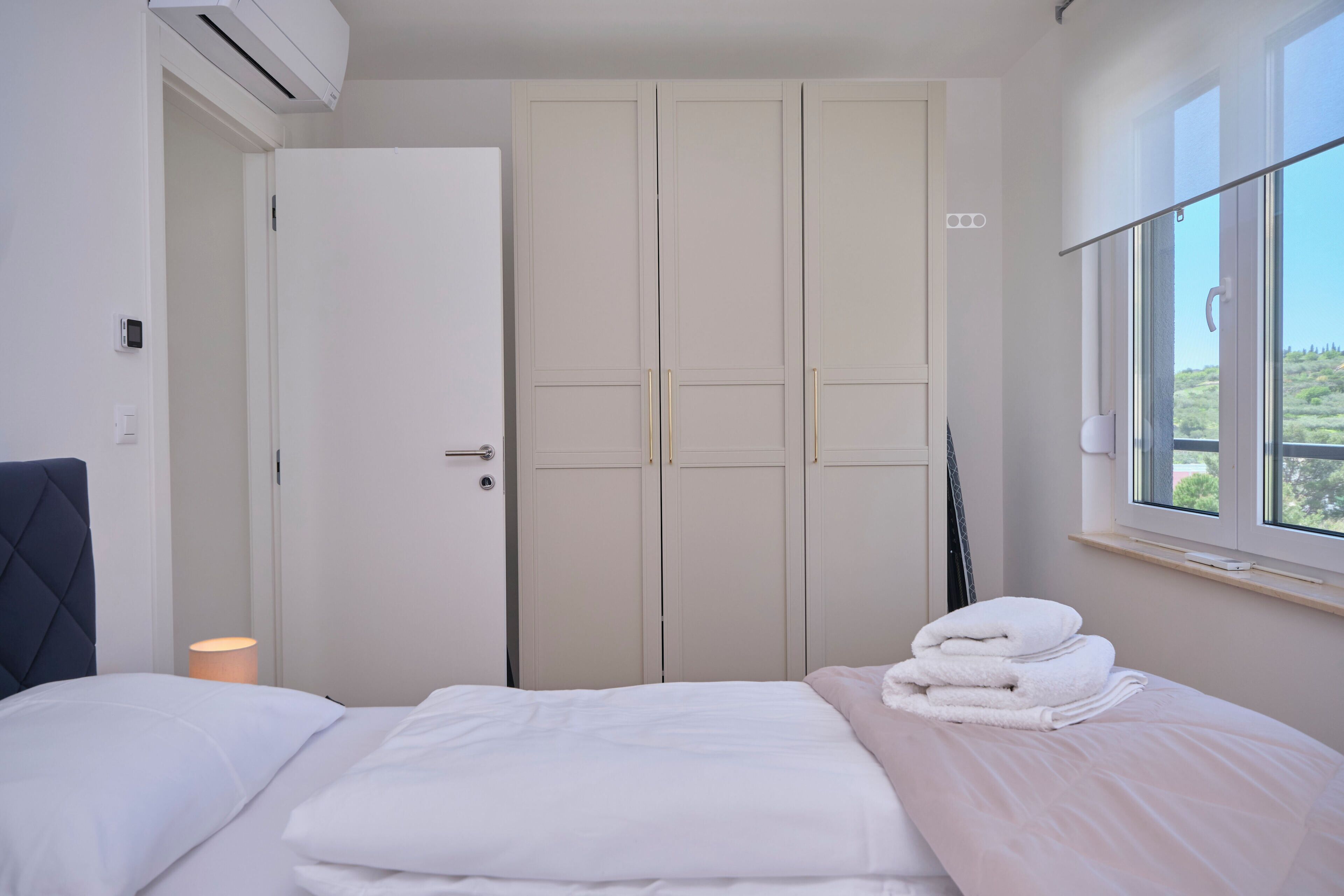A bedroom with a bed, a wardrobe, a window with a view, and an air conditioning unit.