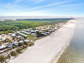 Vista aèria d'una estació turística costanera amb edificis davant de la platja, sorra i una zona boscosa.