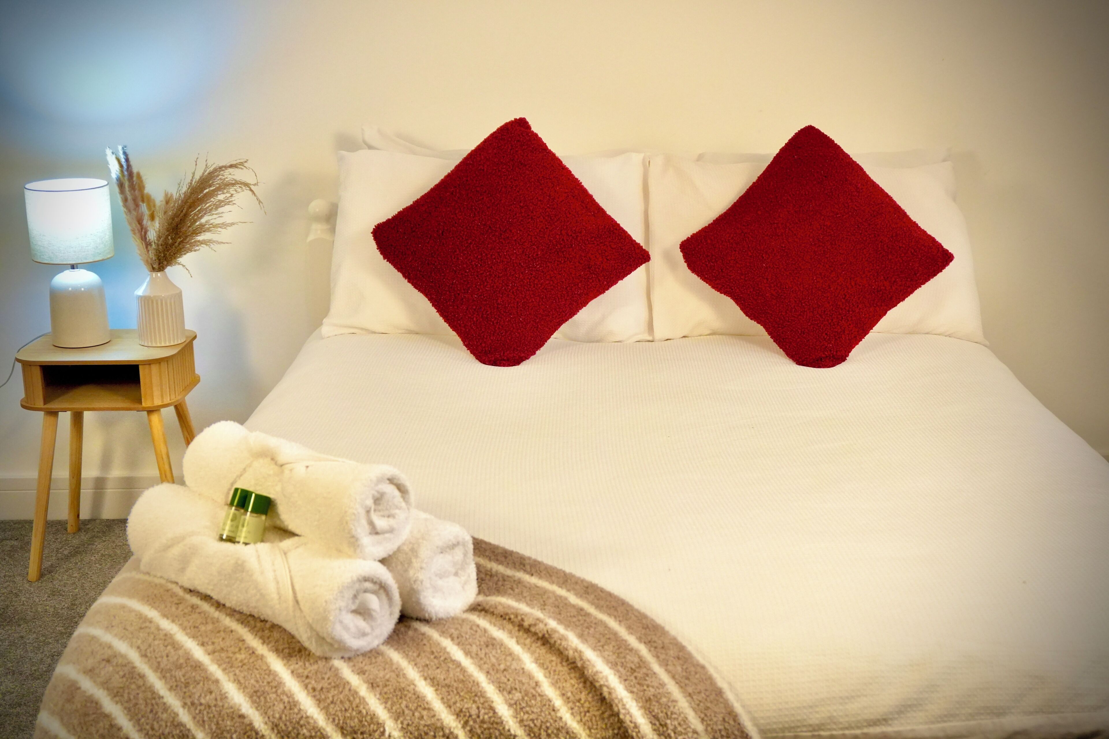 A neatly made bed with red pillows, a bedside lamp, a vase with dried plants, and rolled towels on a striped blanket.