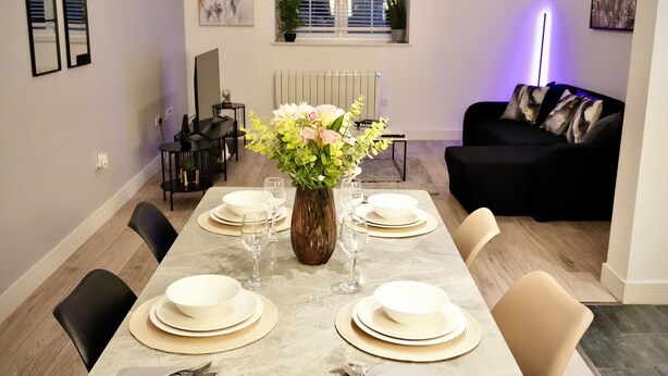 Et spiseområde med et marmorbord dækket til fire personer, omgivet af stole, en vase med blomster og en sort sofa.