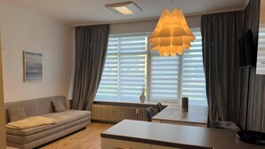 Ein modernes Wohnzimmer mit Sofa, Esstisch, Küchenbereich und einem großen Fenster mit Jalousien.
