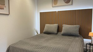Ein ordentlich gemachtes Bett mit gesteppter Bettdecke, zwei Kissen, einem an der Wand hängenden Kunstwerk und einem Kopfteil aus Holzleisten.