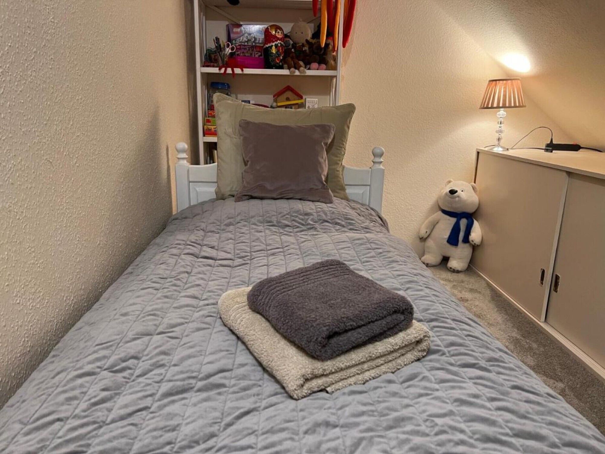 A bedroom with a bed, pillows, a teddy bear, a lamp, and a shelf with toys.