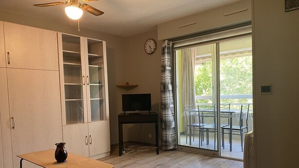 A room with a ceiling fan, a TV on a stand, a dining table with chairs, a cabinet with glass doors, and a sliding glass door leading to an outdoor area. - Charming studio with pool - 2 guests (La Grande-Motte)
