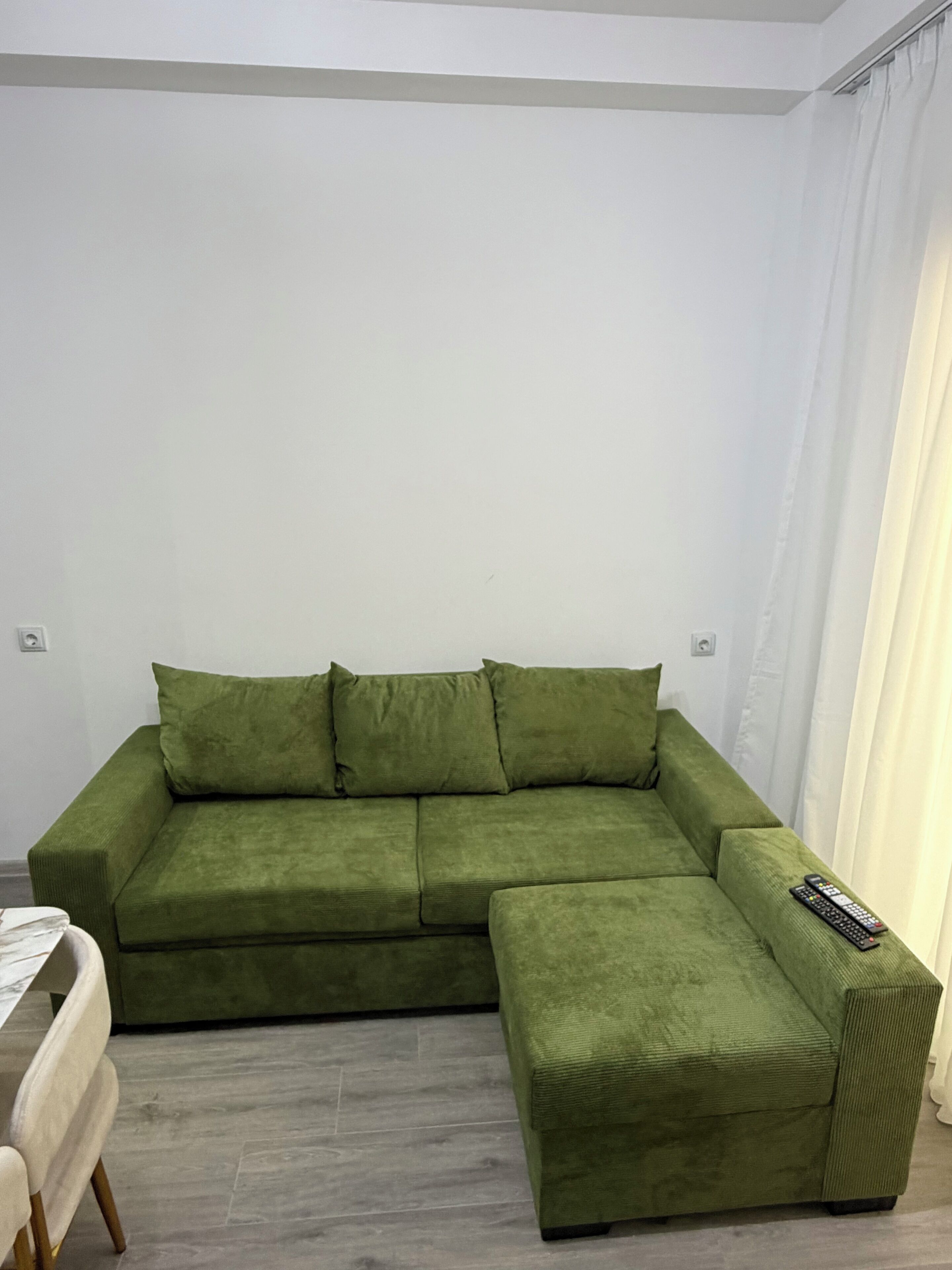 A green sofa with a matching ottoman, a remote control on the ottoman, and a white chair with a table in the foreground.