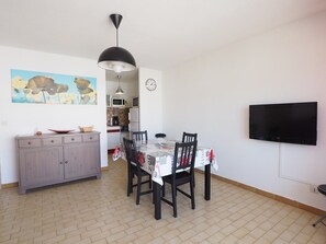 Un comedor con mesa, sillas, un armario, un reloj, un televisor y una cocina al fondo.