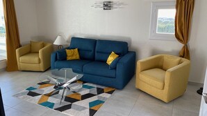 Una sala de estar con un sofá azul, dos sillones amarillos, una mesa de centro de vidrio y una alfombra estampada.