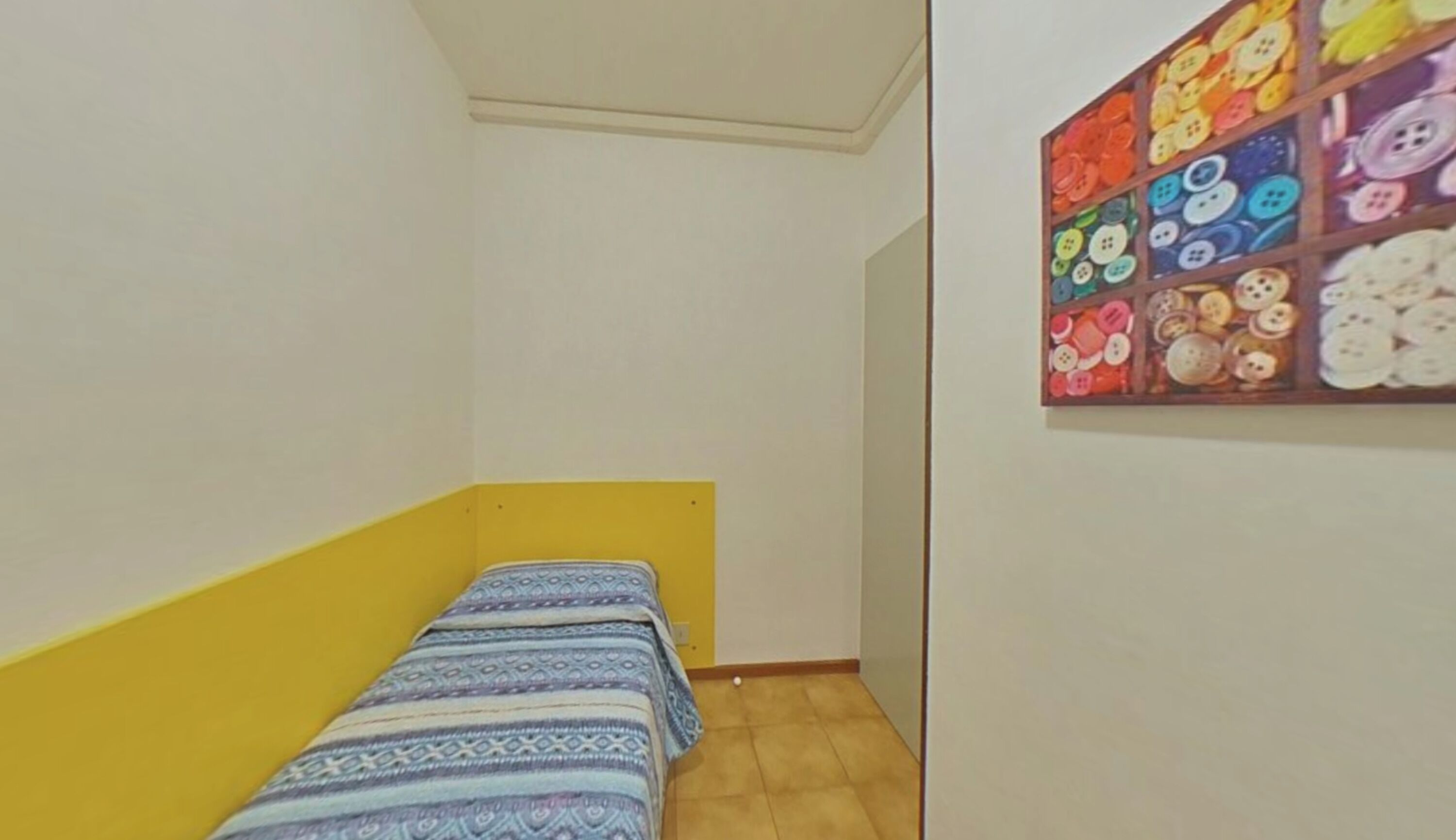 Une petite pièce avec un lit simple, un mur jaune et une décoration murale colorée faite de boutons.