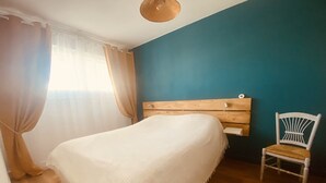 Une chambre à coucher avec un lit, une tête de lit en bois, une chaise blanche et une fenêtre avec des rideaux.