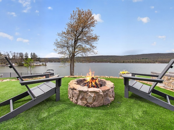 A lakeside deck with a stone fire pit, two lounge chairs, and a view of the lake and hills.