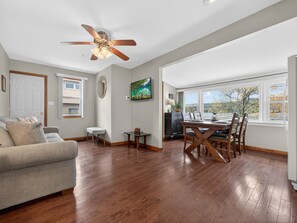 A spacious living room with hardwood flooring, a ceiling fan, a dining area with a table and chairs, and a large window offering a view.