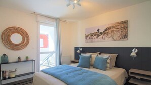 A bedroom with a bed, a bedside table, a wall-mounted lamp, a ceiling fan, and a window with curtains. - Apartment, AC, parking - 4 guests (Le Grau-du-Roi)