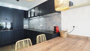 A modern kitchen with dark cabinets, a wooden dining table, wicker pendant lights, and a tiled backsplash. - Apartment, AC, parking - 4 guests (Le Grau-du-Roi)