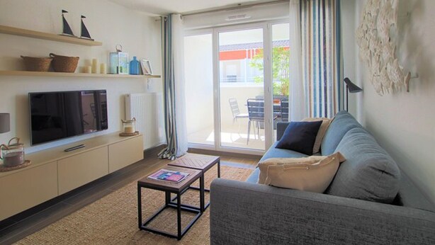 A living room with a sofa, TV, and coffee table, a balcony with a chair, and a view of the outside. - Apartment, AC, parking - 4 guests (Le Grau-du-Roi)