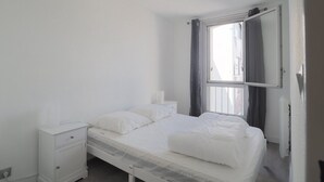 Habitación individual con cama blanca, mesita de noche, ventana con cortinas y armario blanco.