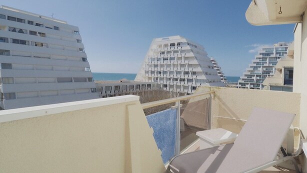 Ein Balkon mit Liegestuhl, Glasgeländer und Blick auf moderne Gebäude sowie das Meer.