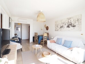 A living room with a sofa, a television, a dining area with chairs, and a large wall art piece. - Sea view apartment, AC, parking - 6 guests (La Grande-Motte)