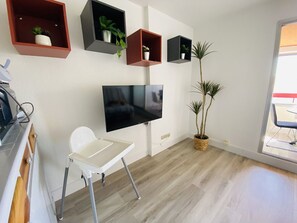 A modern living room with a flat-screen TV, white folding chair, and multiple wall-mounted shelves with potted plants. - Charming apartment with parking - 3 guests (Le Grau-du-Roi)