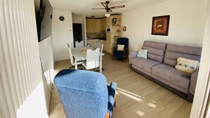 A living room with a sofa, a dining table with chairs, a ceiling fan, and a kitchen area in the background.