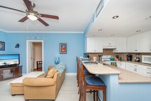 Une cuisine moderne avec des meubles blancs, un coin repas avec une table et des chaises de couleur beige, un ventilateur de plafond et un téléviseur dans le salon.