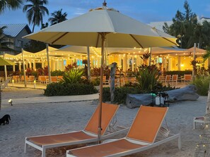 Un espace de détente aménagé au bord de la plage, avec des chaises longues et des parasols, un coin repas avec des tables et des chaises, et une plage de sable bordée de palmiers.