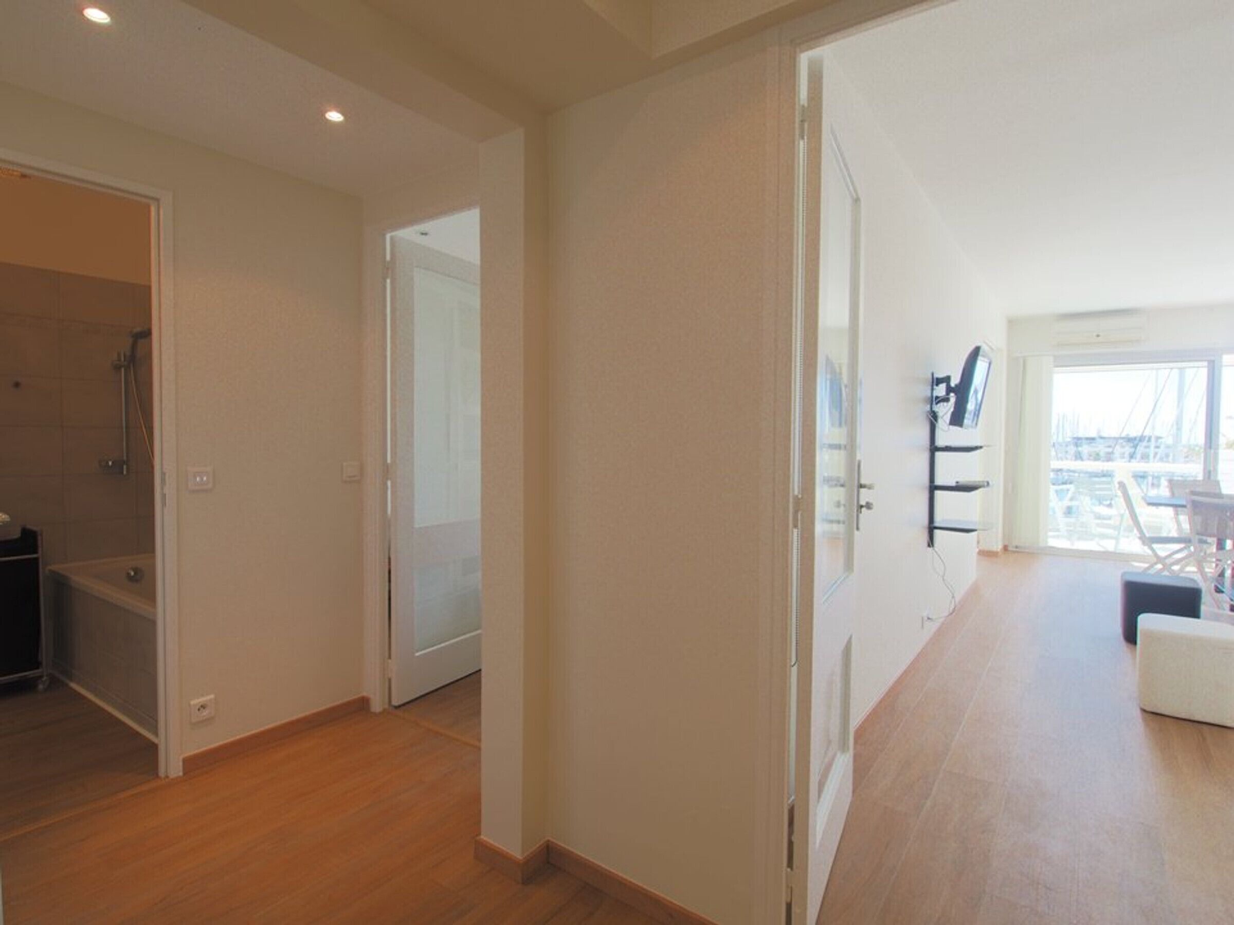 An open-plan living area with a wooden floor, a white door leading to a bathroom, and a view of a marina through a large window.