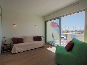 Quarto moderno com sofá, uma poltrona verde, uma mesa pequena e uma janela grande com vista para barcos e água.
