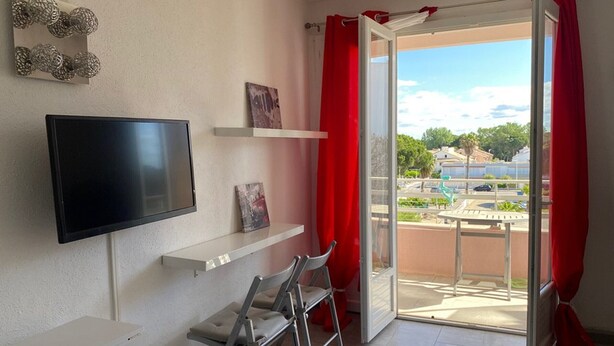 Et værelse med et fladskærms-tv, et lille bord, en stol, en balkon med røde gardiner og udsigt udad.