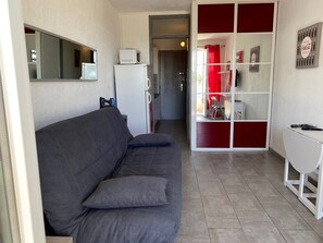 A compact living space with a grey sofa, a white side table, a microwave, and a red-framed shelving unit. - Charming studio near beach, parking - 2 guests (Le Grau-du-Roi)
