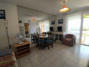 A living room with a dining area, a sofa, a fan, and a TV. - Apartment with pool, parking, lake view - 4 guests (Le Grau-du-Roi)