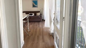 Un pasillo estrecho que conduce a una sala de estar con un sofá y una mesa, un cartel en la pared y un balcón visible a través de un ventanal.
