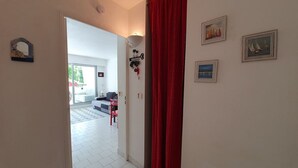 Ein Flur mit rotem Vorhang, gefliestem Boden in Weiß und Blick in einen Wohnbereich mit Sofa und Sessel.