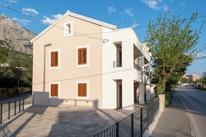 A two-story building with a balcony, a paved courtyard, and a mountain in the background. - Villa Merkur -apartment 1 (Makarska)