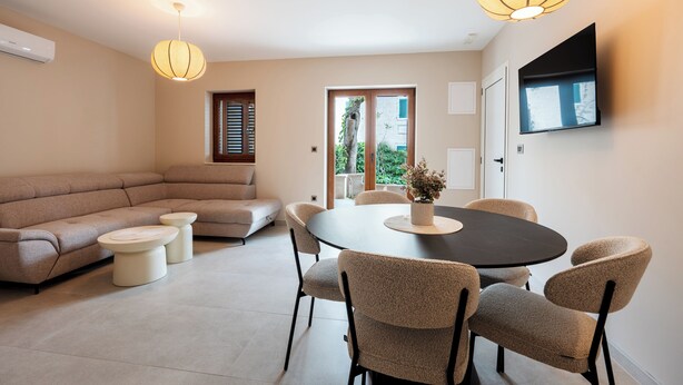 A modern living room with a sectional sofa, a round dining table with chairs, a flat-screen TV mounted on the wall, and a large window with blinds. - Villa Merkur -apartment 1 (Makarska)