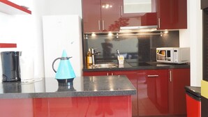 A kitchen with red cabinets, a black countertop, a white refrigerator, a blue kettle, a microwave, and a sink. - Apartment with pool and parking - 4 guests (La Grande-Motte)