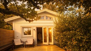 A wooden deck with a bench and chairs, a white building with a door and windows, surrounded by greenery. - Modern guesthouse close to sea, nature, golf, hiking & Kattegat Trail (Särö)