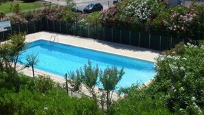 A rectangular swimming pool surrounded by a green fence, with lush greenery and flowering plants in the foreground. - Apartment near the beach - 4 guests (Le Grau-du-Roi)