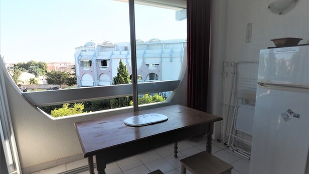A small kitchenette with a refrigerator, a window with a view of buildings, a table, and two stools. - Apartment near the beach - 4 guests (Le Grau-du-Roi)