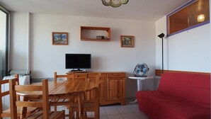 A living room with a wooden dining table, chairs, a red sofa, a television, and artwork on the walls. - Apartment near the beach - 4 guests (Le Grau-du-Roi)