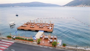 Una plataforma flotante con tumbonas y sombrillas, conectada a un muelle mediante un paseo peatonal.