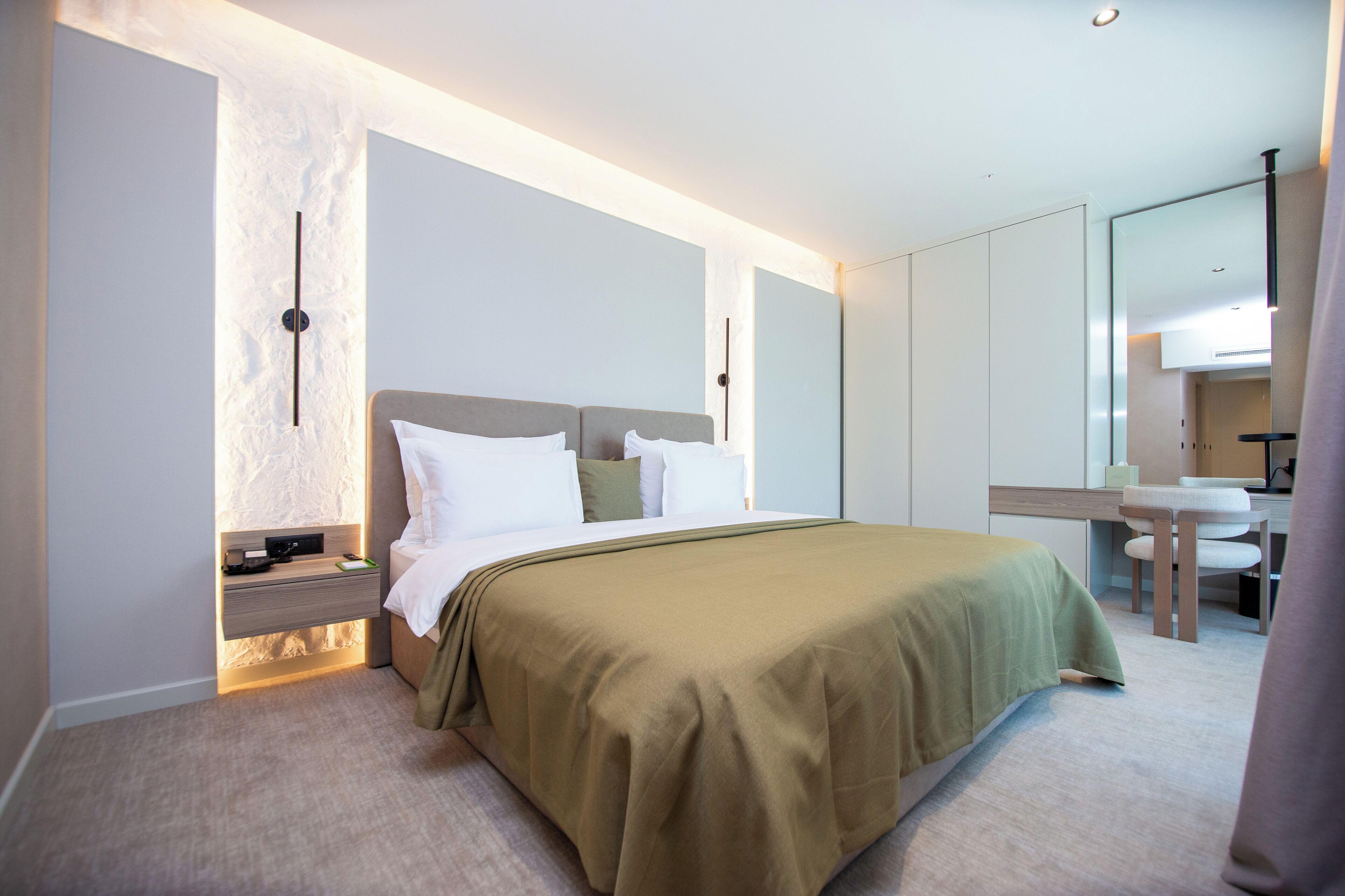 A modern hotel room with a bed, bedside table, and a small desk with a chair.