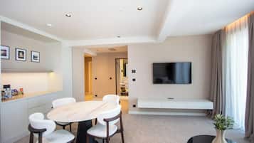 A modern hotel room with a dining area, a flat-screen TV mounted on the wall, a seating bench, and a large window with curtains.